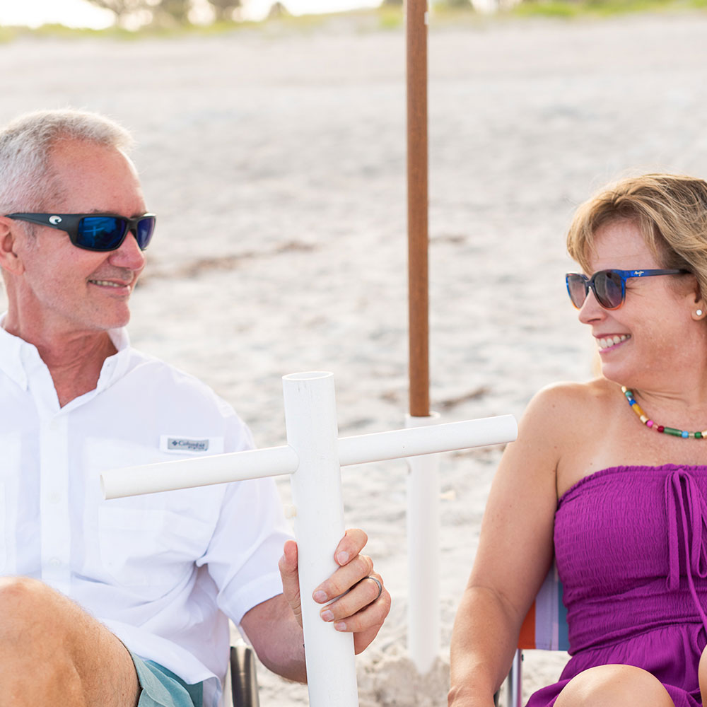 Co-founders Mike & Hope smiling at each other & sitting on beach chairs underneath an umbrella while Mike holds the Suncoast Beach Shade umbrella anchor
