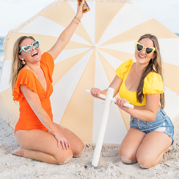 Two women smiling in front of an umbrella as their background as they screw the Suncoast Beach Shade umbrella anchor into the sand at the beach